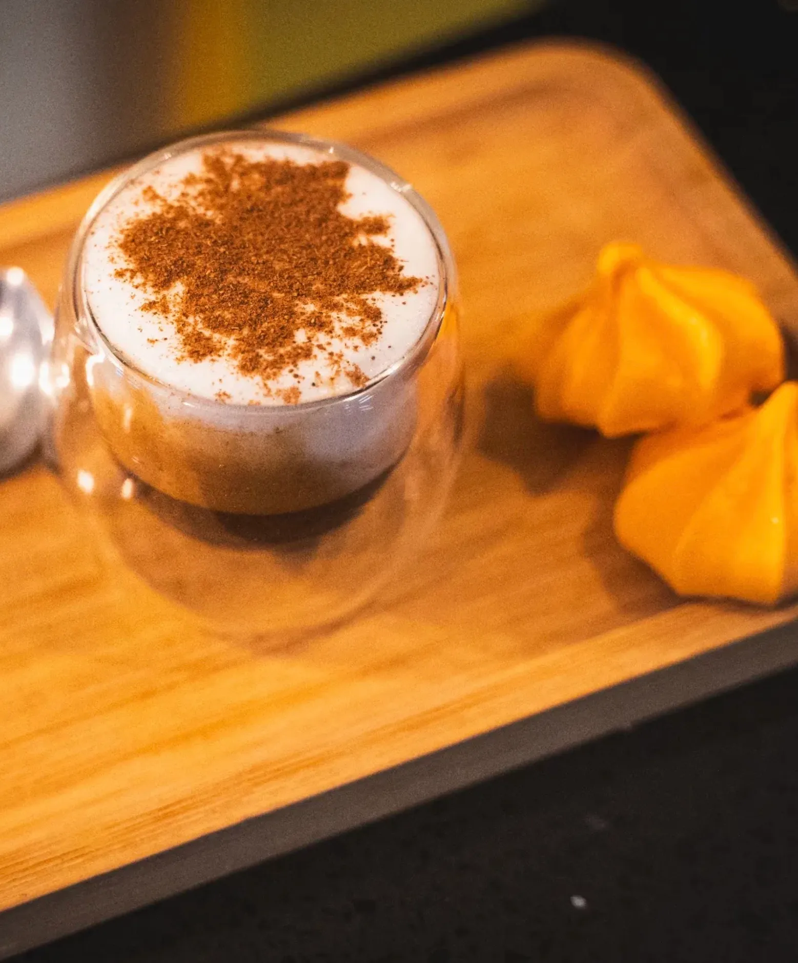 Café con espuma y canela en un vaso de vidrio, sobre una bandeja de madera con cuchara y merengues